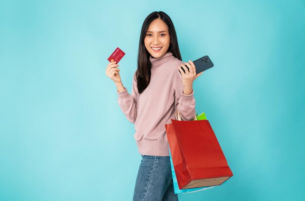 Mooie Aziatische vrouw met papieren boodschappentassen en creditcard met smartphone op blauwe achtergrond.