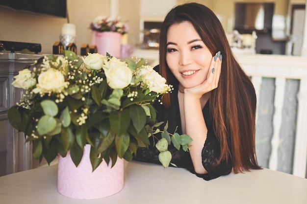 Mooie aziatische vrouw zit bij de tafel in het lichte interieur van de woonkamer en bewondert witte rozenbloemen