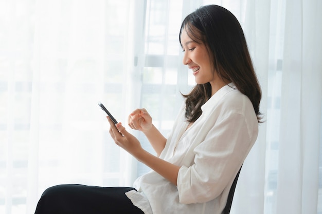 Mooie aziatische vrouw zittend op de stoel en smartphone met typebericht op sociale media vast te houden.