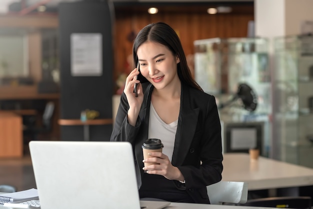 Mooie Aziatische zakenvrouw die aan de telefoon praat en koffie vasthoudt tijdens het chatten met klanten op kantoor.