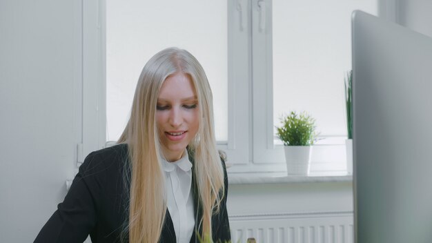 Mooie bedrijfsvrouw die zich op het werk concentreert. Aantrekkelijke jonge blonde vrouw in elegante jas zitten met pen en notebook op bureau met computer en opgewonden kijken naar beeldscherm.