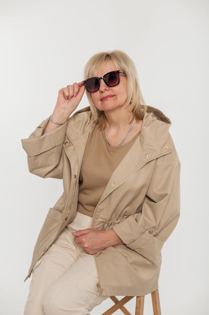 Mooie bejaarde modieuze blonde vrouw in stijlvolle beige kleding met een jas zet een zonnebril op en zit op een stoel op een witte achtergrond