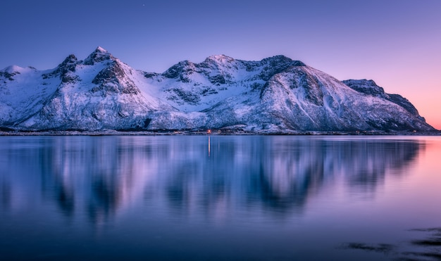 Mooie besneeuwde bergen en kleurrijke hemel weerspiegeld in het water in de schemering. winterlandschap met zee, besneeuwde rotsen, paarse hemel, reflectie, bij zonsondergang.