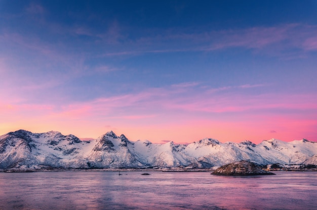 Mooie besneeuwde bergen en kleurrijke hemel weerspiegeld in het water in de schemering. Winterlandschap met zee, besneeuwde rotsen, paarse hemel, reflectie, bij zonsondergang.