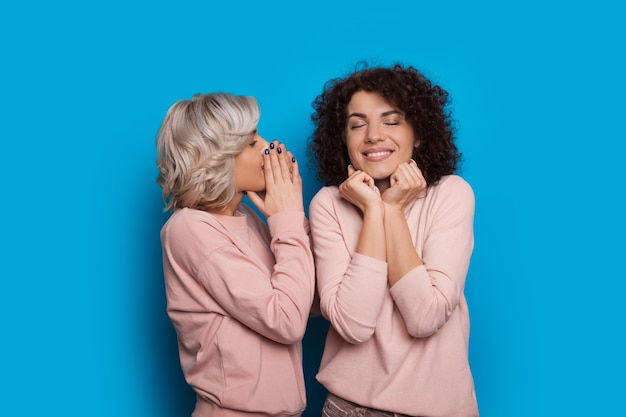 Mooie blanke dame met krullend haar luistert naar haar blonde vriend en geniet van moment terwijl poseren op een blauwe achtergrond