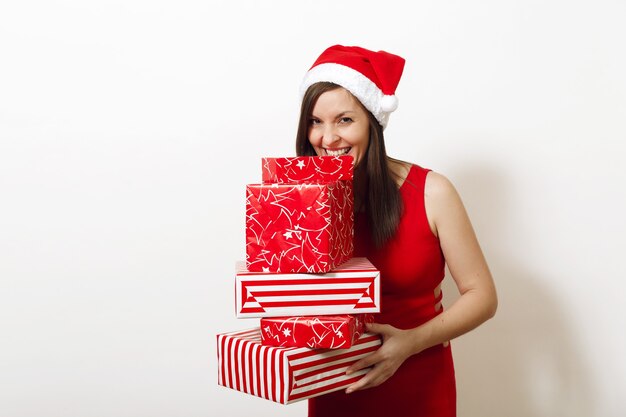 Mooie blanke jonge gelukkige vrouw met charmante glimlach gekleed in rode jurk en kerstmuts met geschenkdozen op witte achtergrond. Santa meisje met heden geïsoleerd. Nieuwjaarsvakantie 2018 concept