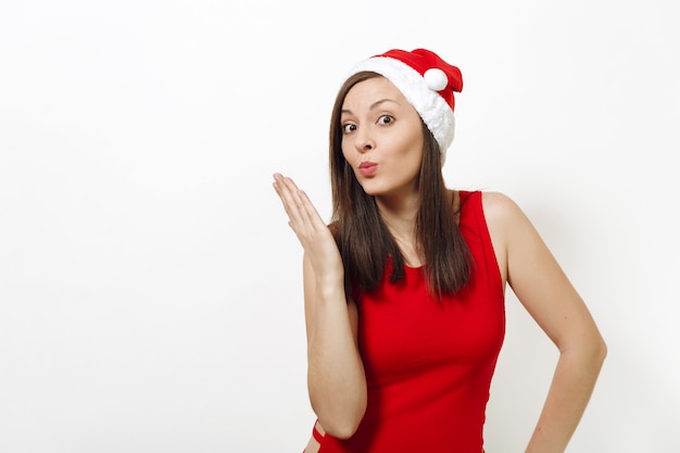 Mooie blanke jonge gelukkige vrouw met een gezonde huid en charmante glimlach met een rode jurk en een kerstmuts die een luchtkus verzendt op een witte achtergrond. Santa meisje geïsoleerd. Nieuwjaarsvakantie 2018 concept.