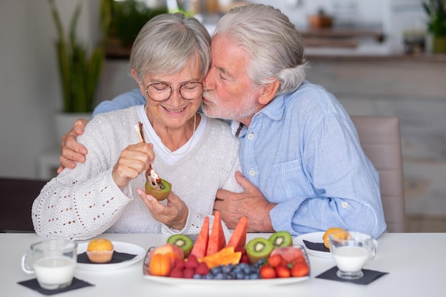 Mooie blanke senior man die zijn vrouw kust terwijl hij thuis ontbijt met verse seizoensfruitmelk en cupcake gezond eetconcept