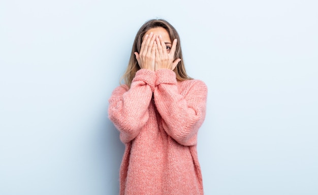 Mooie blanke vrouw die haar gezicht bedekt met handen, tussen de vingers gluurt met een verbaasde uitdrukking en opzij kijkt