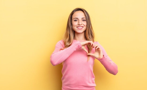 Mooie blanke vrouw die lacht en zich gelukkig, schattig, romantisch en verliefd voelt, en met beide handen hartvorm maakt