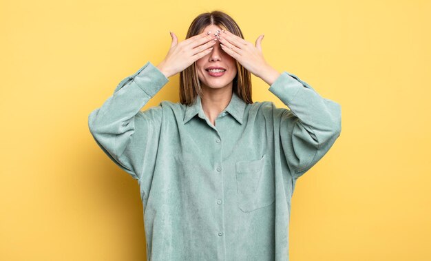 Mooie blanke vrouw die lacht en zich gelukkig voelt, de ogen met beide handen bedekt en wacht op een ongelooflijke verrassing