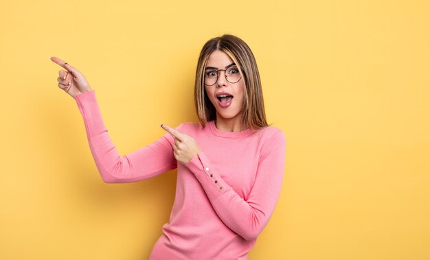 Mooie blanke vrouw die zich blij en verrast voelt, glimlacht met een geschokte uitdrukking en naar de zijkant wijst