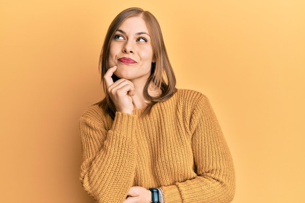 Mooie blanke vrouw, gekleed in een casual wintertrui, denkend geconcentreerd over twijfel met de vinger op de kin en opkijkend, zich afvragend