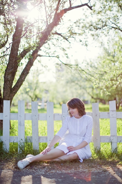 Mooie blanke vrouw in witte jurk loopt in de lentebloesemtuin in de buurt van een witte houten omheining