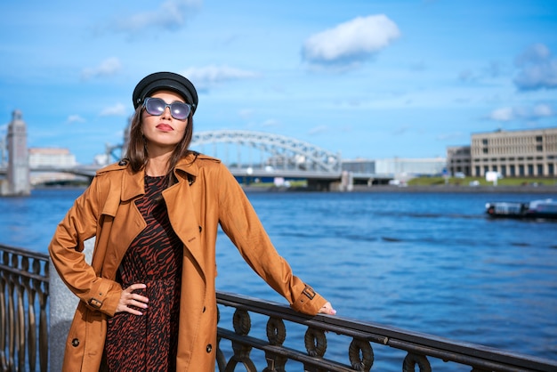 Mooie blanke vrouw in zonnebril in een zwarte pet en jas poseren terwijl ze op de dijk staat op een zonnige dag van blauwe lucht en stadsgezicht