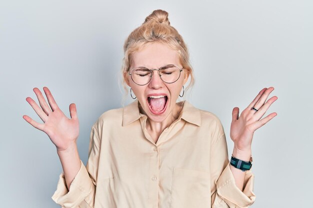 Mooie blanke vrouw met blond haar met een casual look en een bril die gek en gek viert voor succes met opgeheven armen en gesloten ogen schreeuwend opgewonden winnaarconcept