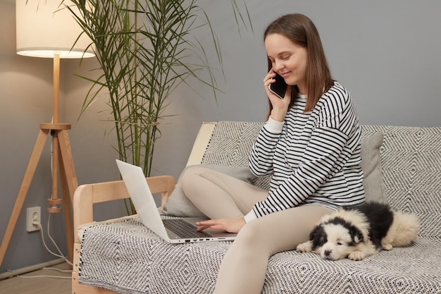 Mooie blanke vrouw met een gestreepte hemd die op de bank zit met haar puppy die aan het bellen is.