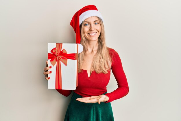 Mooie blanke vrouw met kerstmuts en met een geschenk dat er positief en gelukkig uitziet en glimlacht met een zelfverzekerde glimlach die tanden laat zien