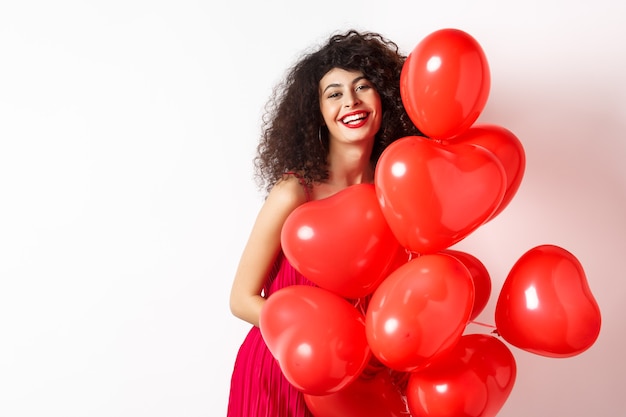 Mooie blanke vrouw met krullend haar, date jurk dragen, romantische rode harten ballonnen houden en lachen op Valentijnsdag, gelukkig permanent op witte achtergrond.