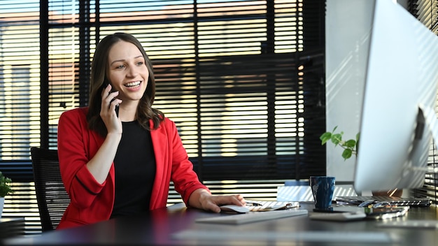 Mooie blanke zakenvrouw die een telefoongesprek heeft met de klant en naar het computerscherm kijkt op het bureau