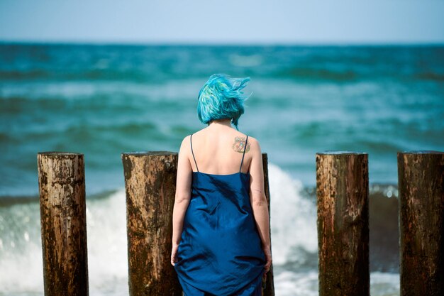 Mooie blauwharige vrouw performance artiest in donkerblauwe jurk staande op strand met verfborstel, achteraanzicht. Hedendaagse abstracte kunst, danskunst in de natuur, moderne performancekunst buiten