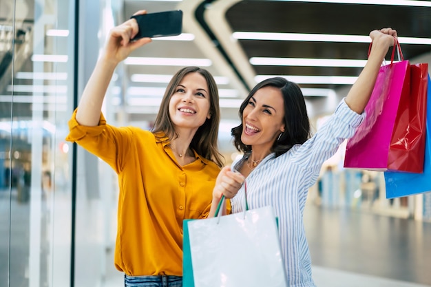 Mooie blij en opgewonden jonge vriendinnen met papieren zakken en slimme telefoon lopen rond het winkelcentrum