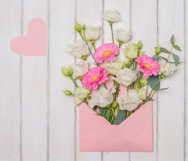 Mooie bloemen in roze envelop en roze valentijnskaarthart op witte houten achtergrond