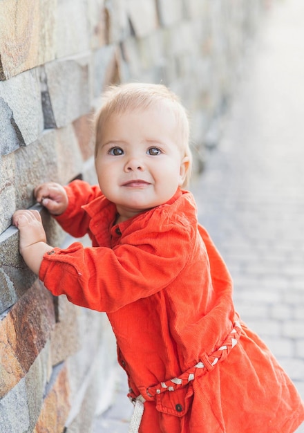 mooie blonde baby in een rode jurk staat in de buurt van een stenen muur