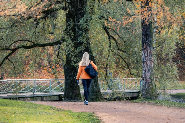 Mooie blonde in een oranje jasje loopt door een herfst steegje. Het uitzicht vanaf de achterkant.