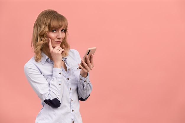 Mooie blonde in een shirt met een telefoon in haar handen, mysterieus glimlachend op een roze achtergrond, het concept van een meisje en een telefoon, kopieer ruimte.