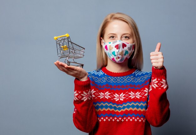 Mooie blonde in Kerstmissweater met boodschappenwagentje op grijs