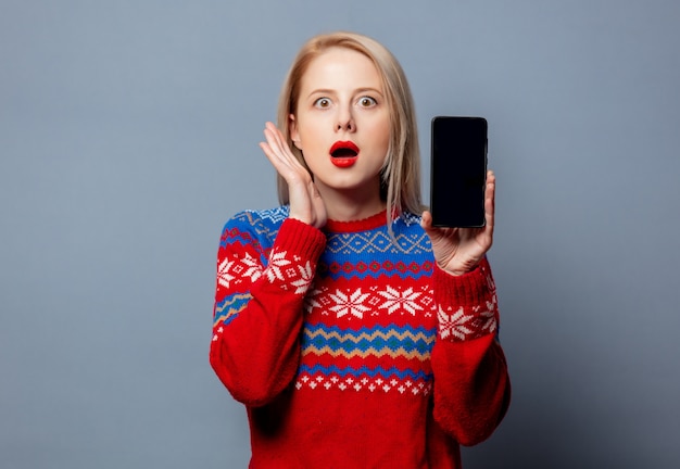 Mooie blonde in Kerstmissweater met mobiele telefoon op grijs