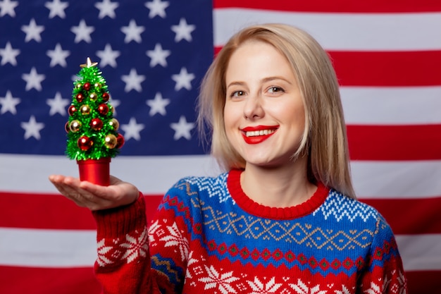 Mooie blonde in kersttrui houdt een boom op de vlag van de VS.