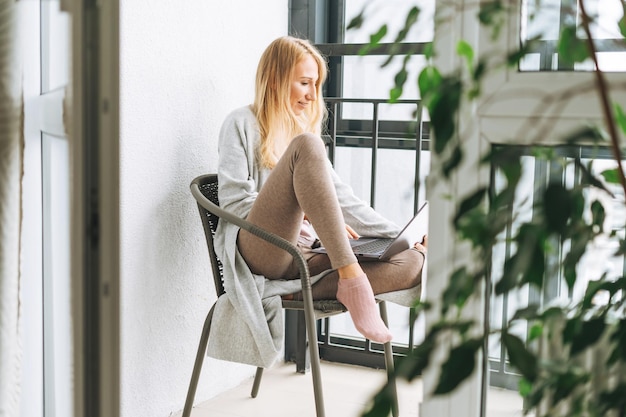 Mooie blonde jonge vrouw freelancer in huiskleding met behulp van laptop op het balkon