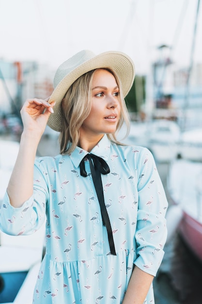 Mooie blonde jonge vrouw in blauwe jurk en strohoed op de pier bij zonsondergang