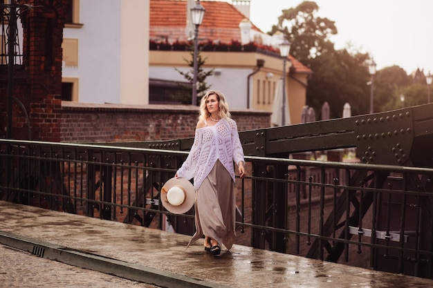 Mooie blonde jonge vrouw met jurk en strohoed die geniet van zonsopgang aan de kust in de oude Europese stad Mode en stijl Zomerreizen