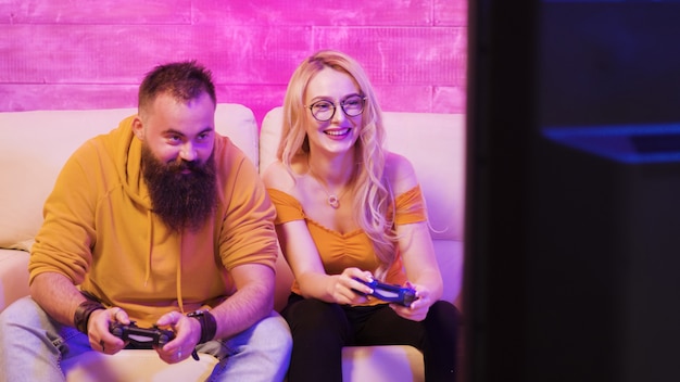 Mooie blonde meid die lacht terwijl ze videogames speelt met haar vriendje met behulp van draadloze controllers op een grootbeeld-tv