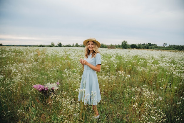 mooie blonde meisje in een veld van madeliefjes. vrouw in een blauwe jurk in een veld van witte bloemen. meisje met een boeket van madeliefjes. zomer tedere foto in het dorp. wilde bloemen. meisje in een strooien hoed