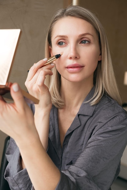 Mooie blonde vrouw die in de spiegel kijkt maakt make-up met concealer en corrector foundation