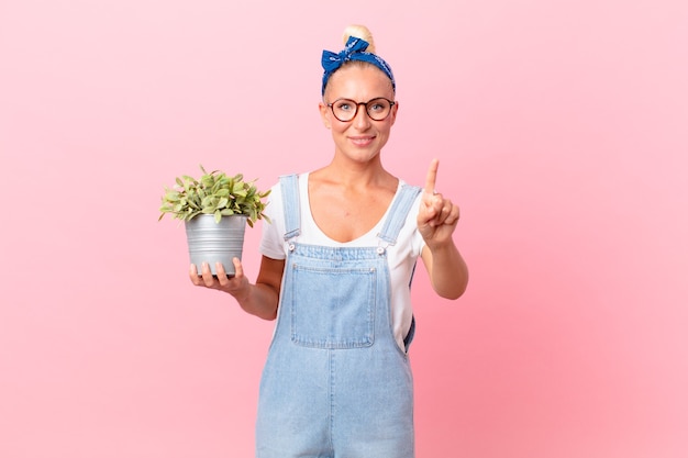 Mooie blonde vrouw die lacht en er vriendelijk uitziet, nummer één toont en een plant vasthoudt