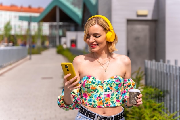 Mooie blonde vrouw die naar muziek luistert in de stad met gele koptelefoons en de telefoonlevensstijl van een model in de stad