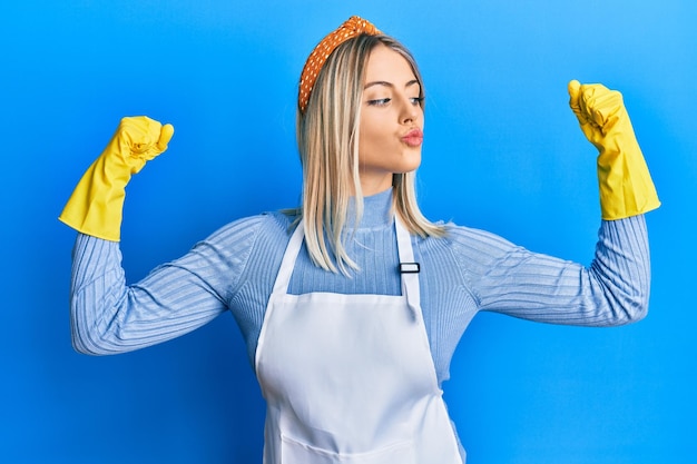 Mooie blonde vrouw die schonere schort en handschoenen draagt die wapenspieren tonen die trots fitnessconcept glimlachen