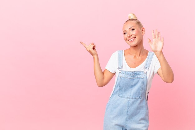 Mooie blonde vrouw die vrolijk lacht, met de hand zwaait, je verwelkomt en begroet