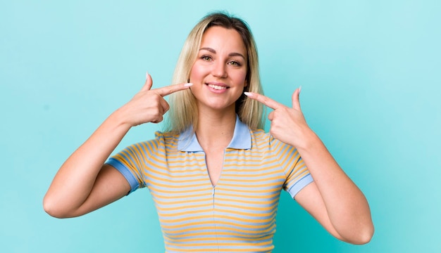 Mooie blonde vrouw die zelfverzekerd glimlacht en wijst naar haar eigen brede glimlach positieve ontspannen tevreden houding