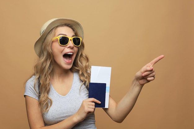 Mooie blonde vrouw die zomerkleren draagt die met paspoort met kaartjes over beige achtergrond stellen.