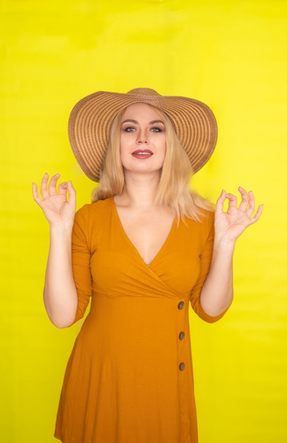 Mooie blonde vrouw in bruine hoed en donker gele jurk poseren over gele muur. Mode zomerlook