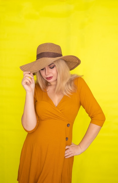Mooie blonde vrouw in bruine hoed en donker gele jurk poseren over gele muur. Mode zomerlook