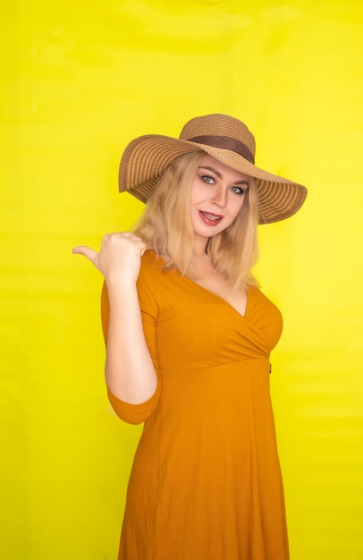 Mooie blonde vrouw in bruine hoed en donker gele jurk poseren over gele muur. Mode zomerlook