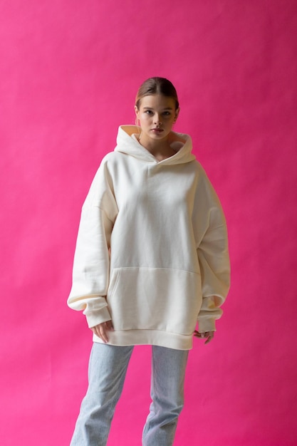 Mooie blonde vrouw in een witte hoodie en blauwe spijkerbroek die op een roze achtergrond poseert