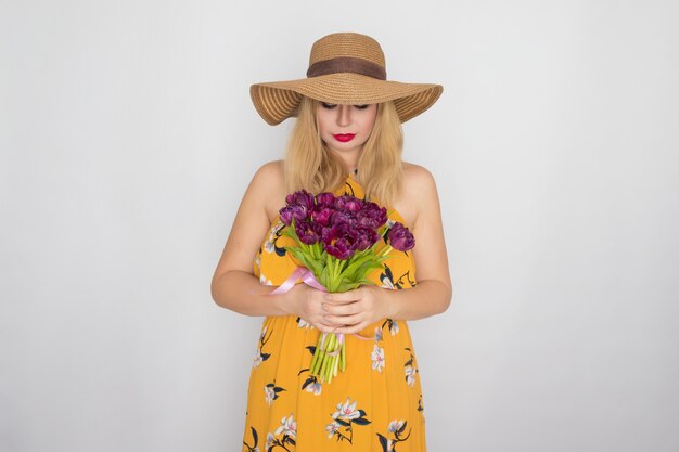 Mooie blonde vrouw in gele bloemen jurk en strooien hoed met boeket paarse tulpen